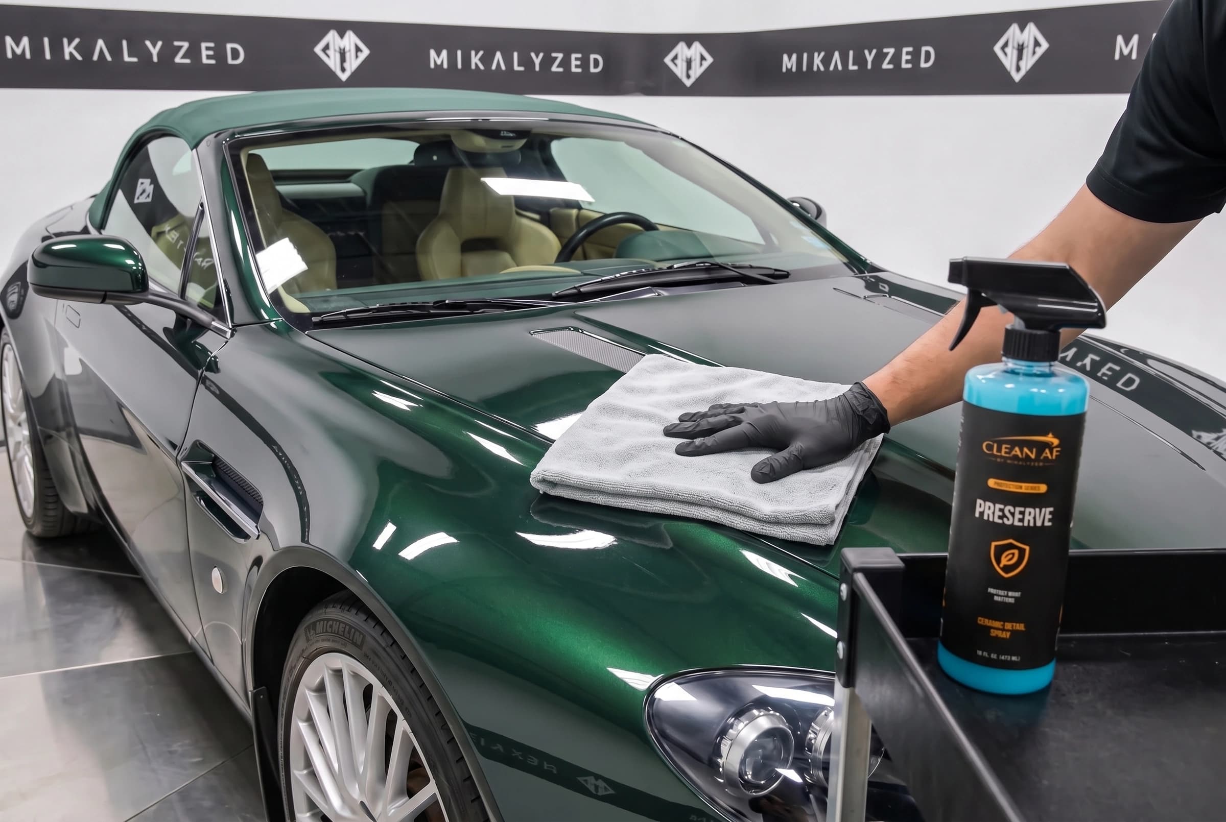 A gray microfiber towel spreading PRESERVE across the green Aston Martin hood