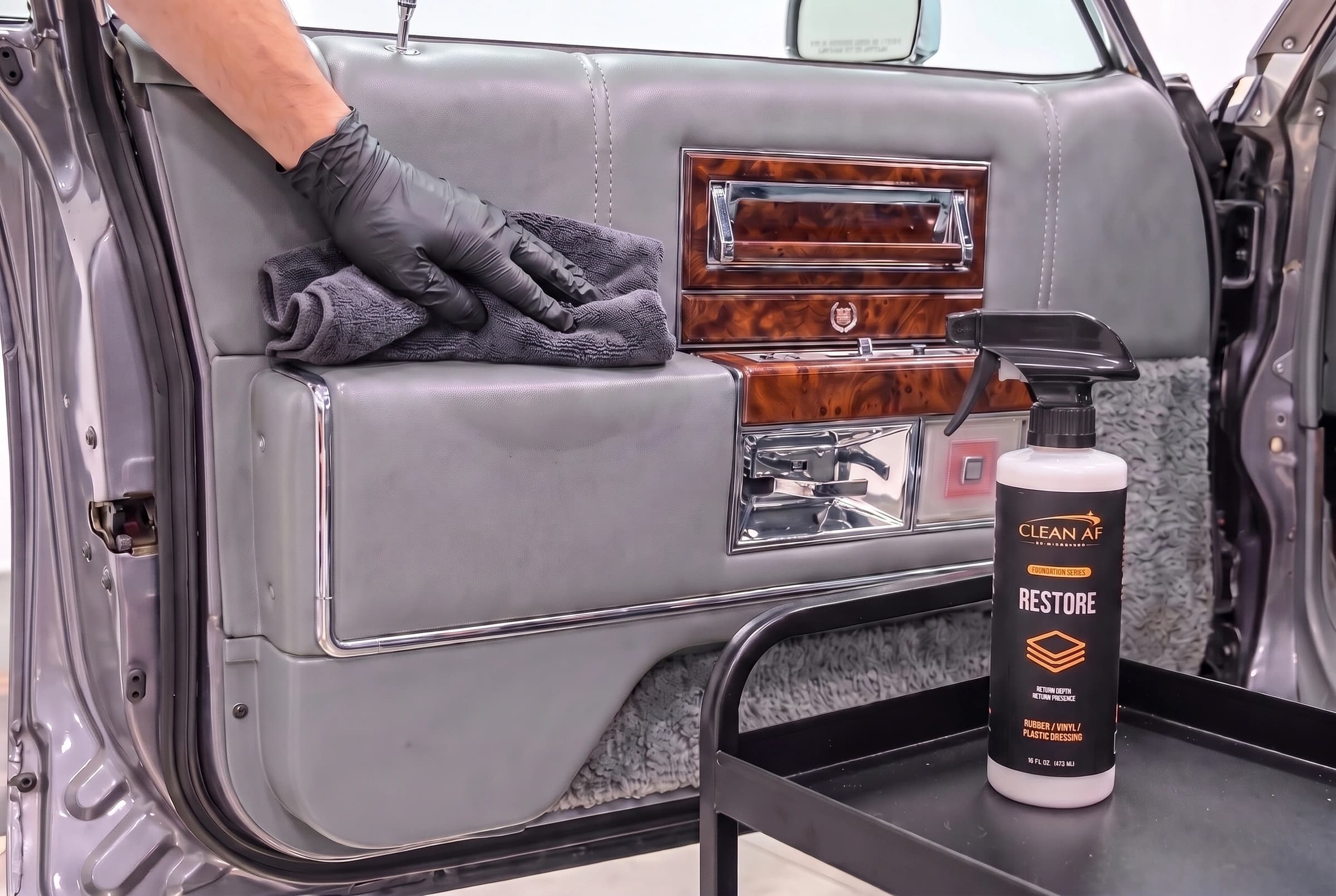 A gloved hand wiping the gray Cadillac vinyl armrest with a folded microfiber towel beside RESTORE